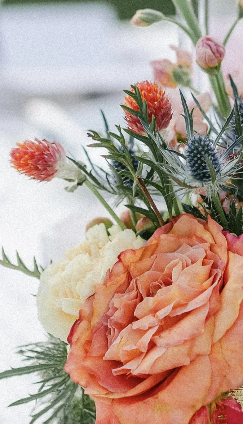Close-up of a colorful flower arrangement with peach roses, small orange and pink flowers, and blue thistle.