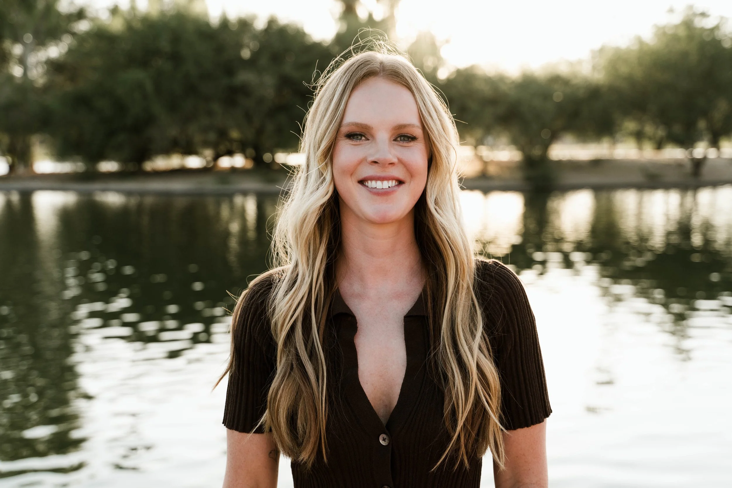 A smiling woman with long blonde hair stands outdoors near a body of water, with trees and the setting sun in the background.
