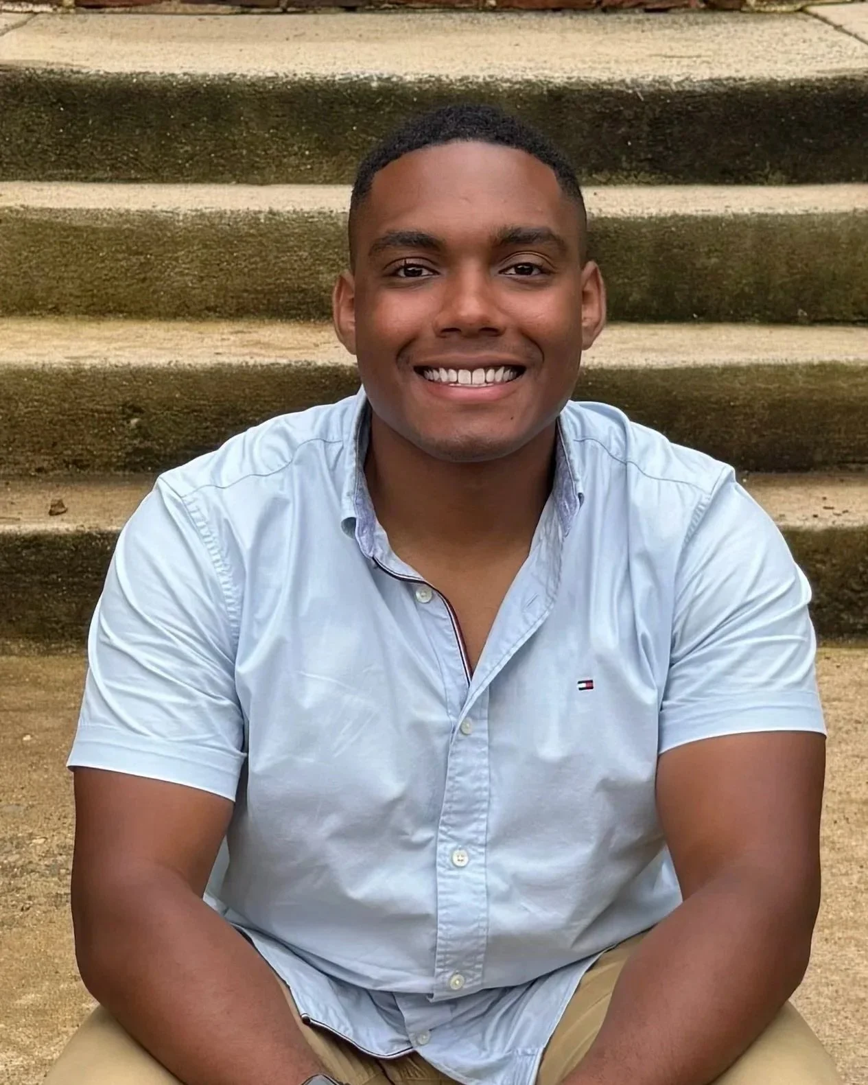 Young man with short black hair smiling, wearing a light blue button-up shirt and khaki pants, sitting outdoors in front of concrete steps.