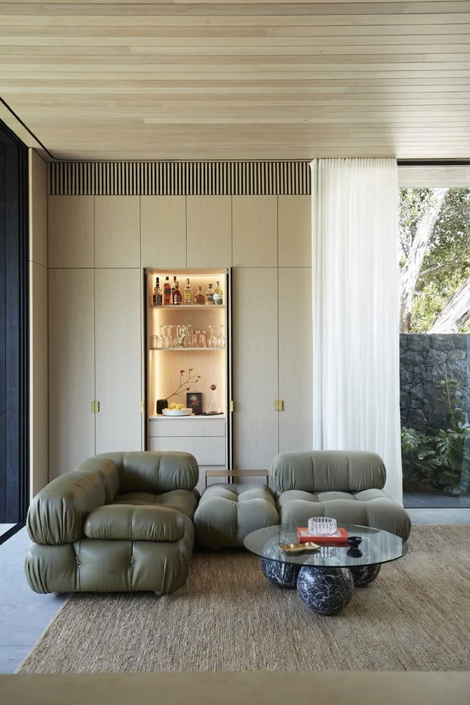 Modern living room with two green leather armchairs, a glass coffee table, a tan rug, beige wall panels, a built-in illuminated liquor shelf, white curtains, and outdoor view of trees and rocks.