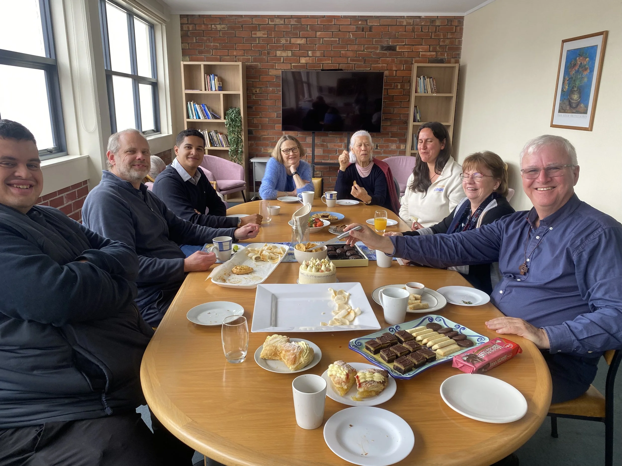 People sitting around a dining table with desserts, coffee, and orange juice, in a room with brick wall, bookshelf, and a mountain landscape painting.