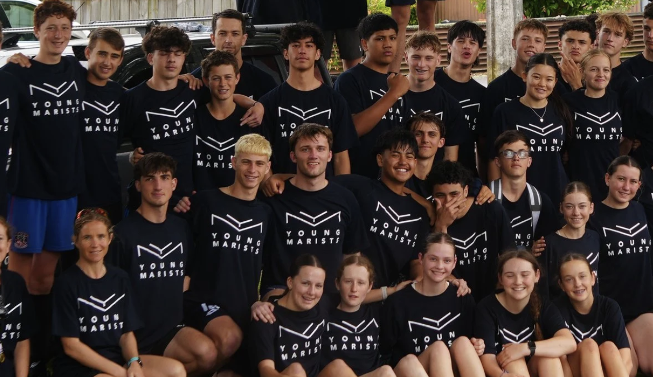 Group photo of Young Marists wearing matching black t-shirts with 'Young Marists' logo, outdoors, some standing, others kneeling, smiling and posing for the camera.