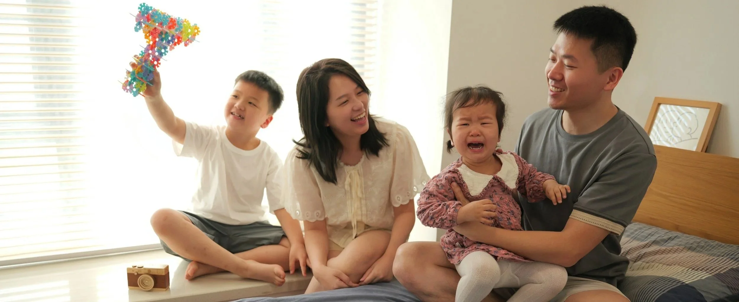 Marist Families with two children, one of whom is crying, sitting on a bed in a bright room with blinds. One child is holding a colorful paper arrow, and the parents are smiling.