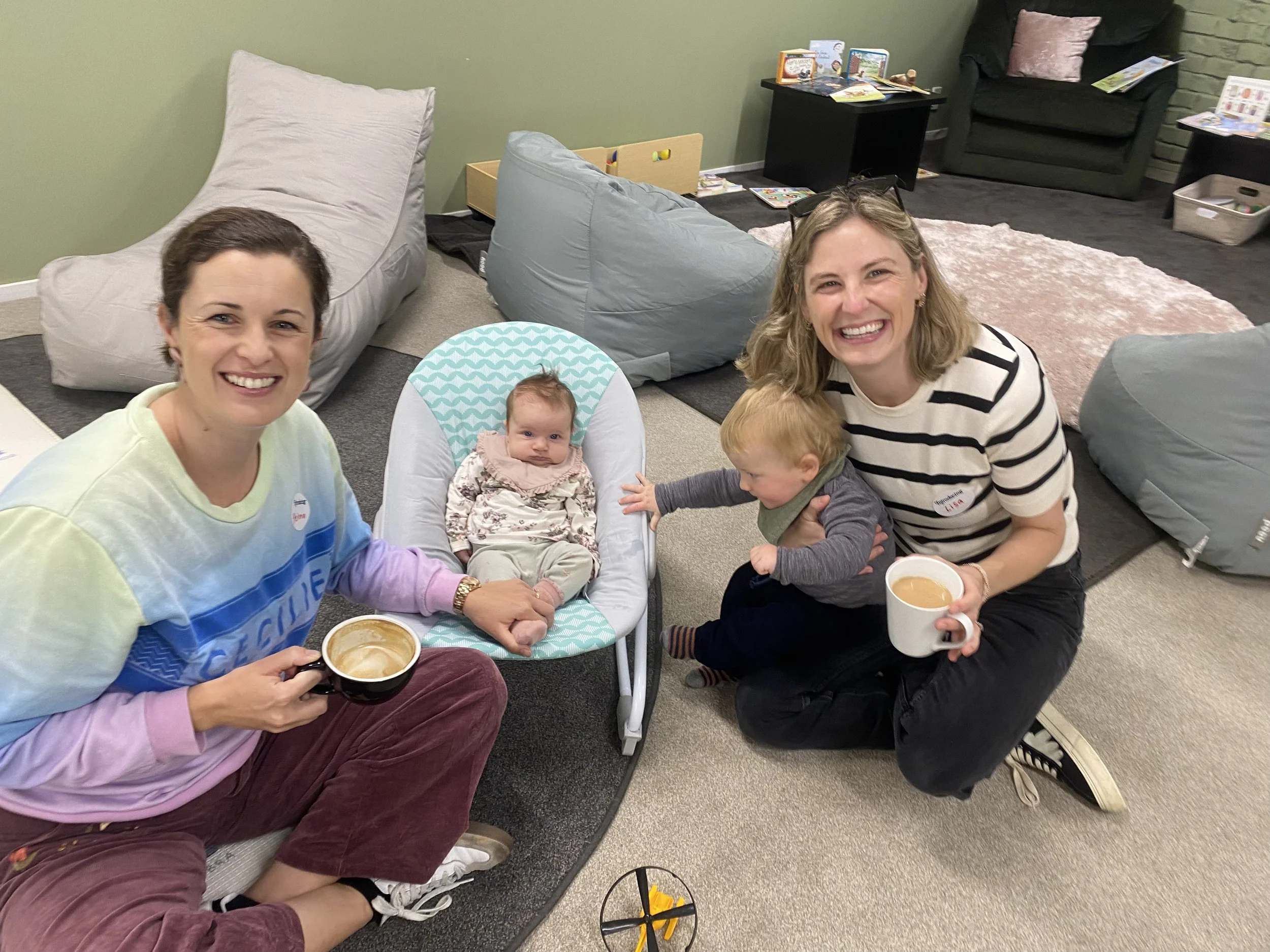 Marist Families and two women, a baby in a high chair, and a young child in a room with bean bag chairs, books, and a pink rug, at marist families cuppa and play group