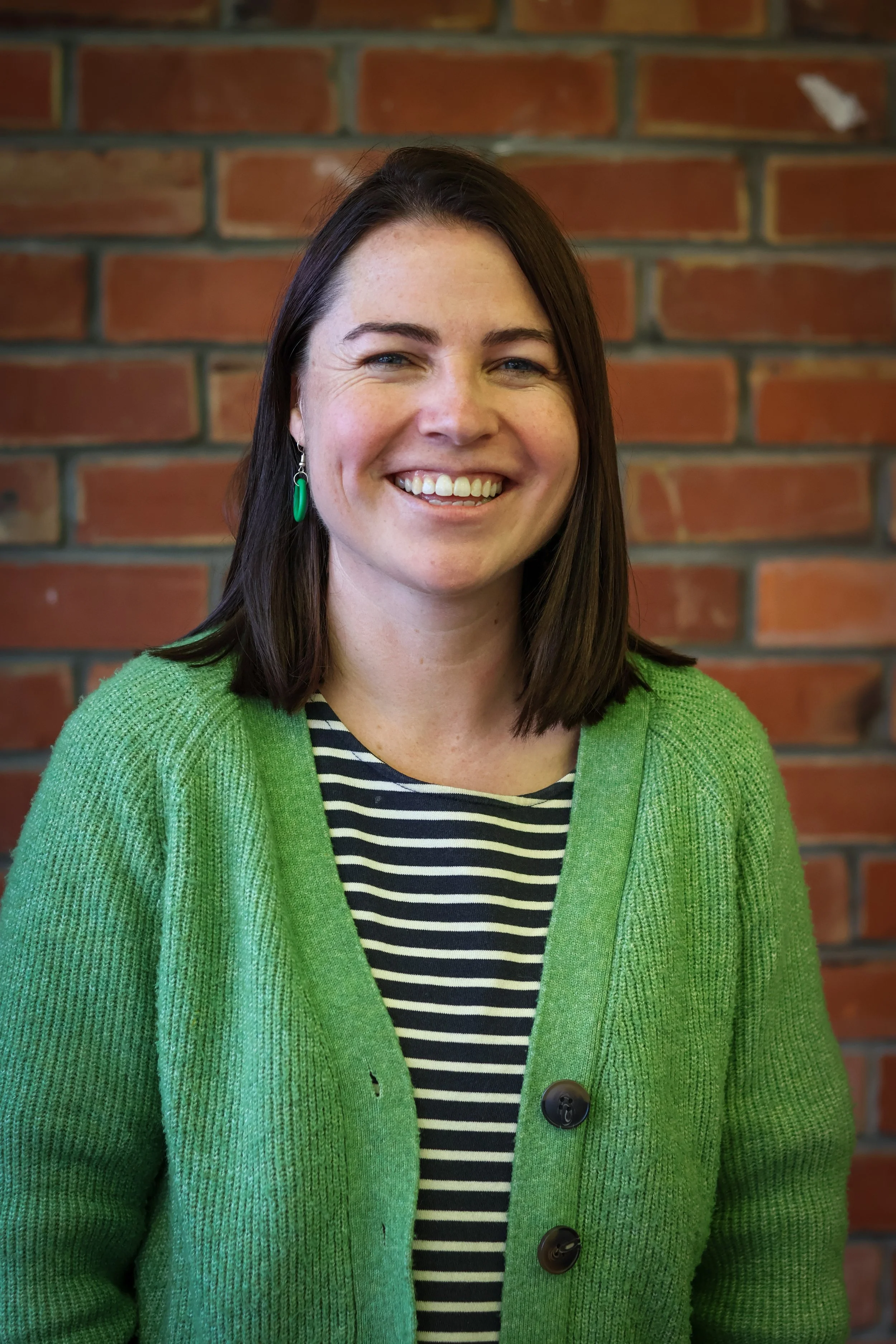 A woman with shoulder-length brown hair and blue eyes smiling, wearing a green cardigan over a black and white striped shirt, and green earrings, standing in front of a red brick wall.