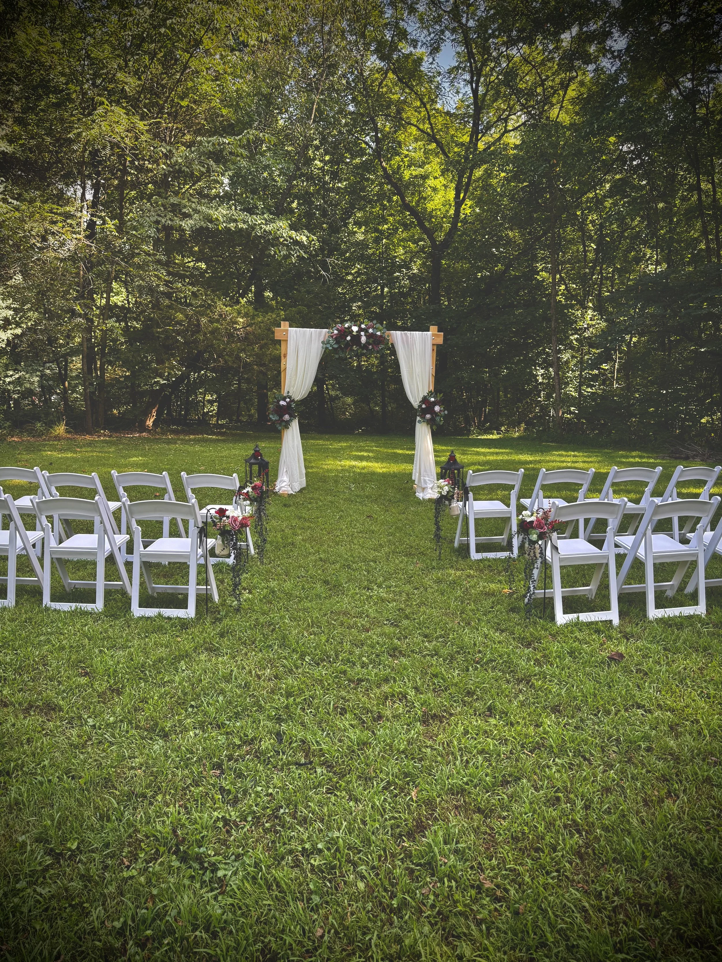 Outdoor wedding ceremony setup with white chairs arranged on green grass facing an altar decorated with white curtains and floral arrangements, surrounded by trees in a wooded area.
