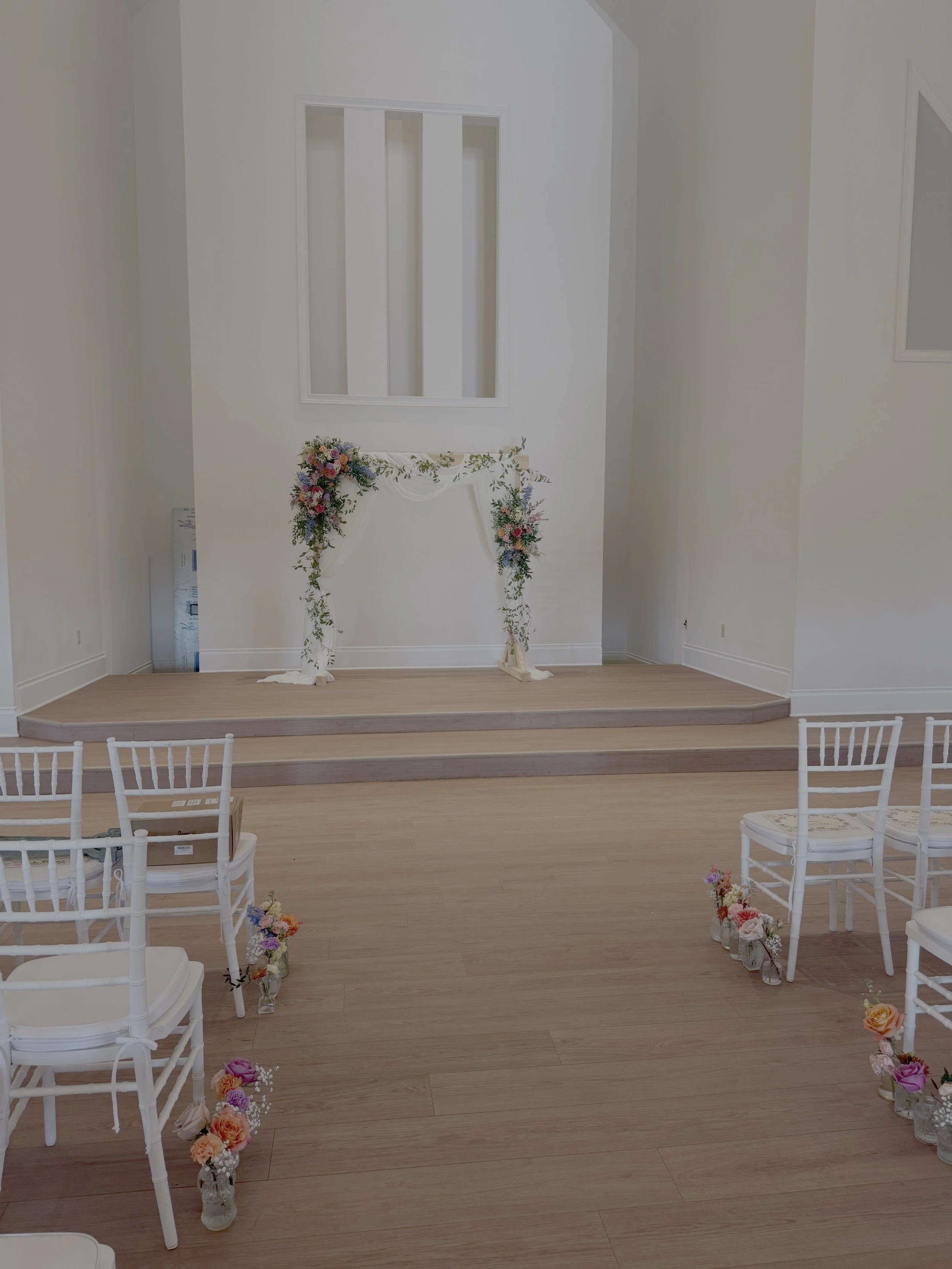 An empty wedding ceremony space with white chairs decorated with flowers along the aisle, a small stage with a floral arch, and a plain white wall backdrop.