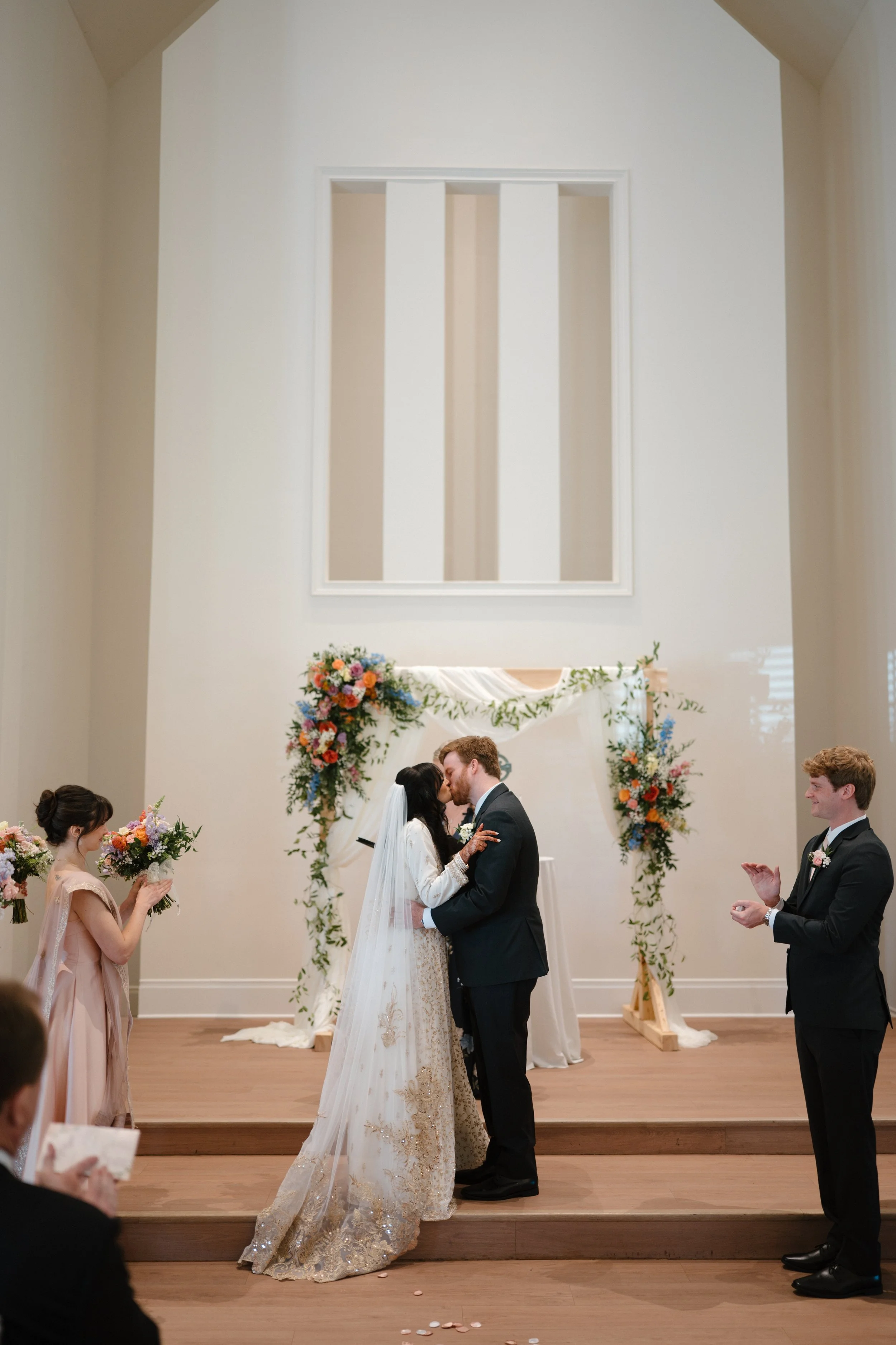 A wedding ceremony taking place inside a church. The bride and groom are kissing at the altar, surrounded by floral arrangements. Bridesmaids and groomsmen are standing nearby, with bridesmaids holding bouquets and groomsmen clapping.
