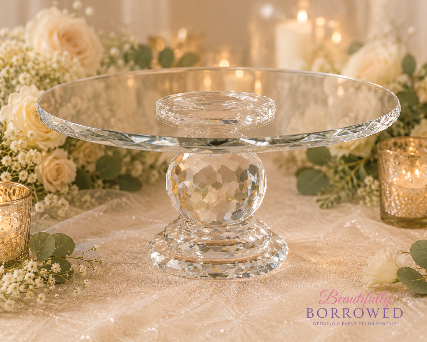 Elegant glass cake stand with a diamond-patterned base, surrounded by white flowers and candle lanterns for wedding decor.