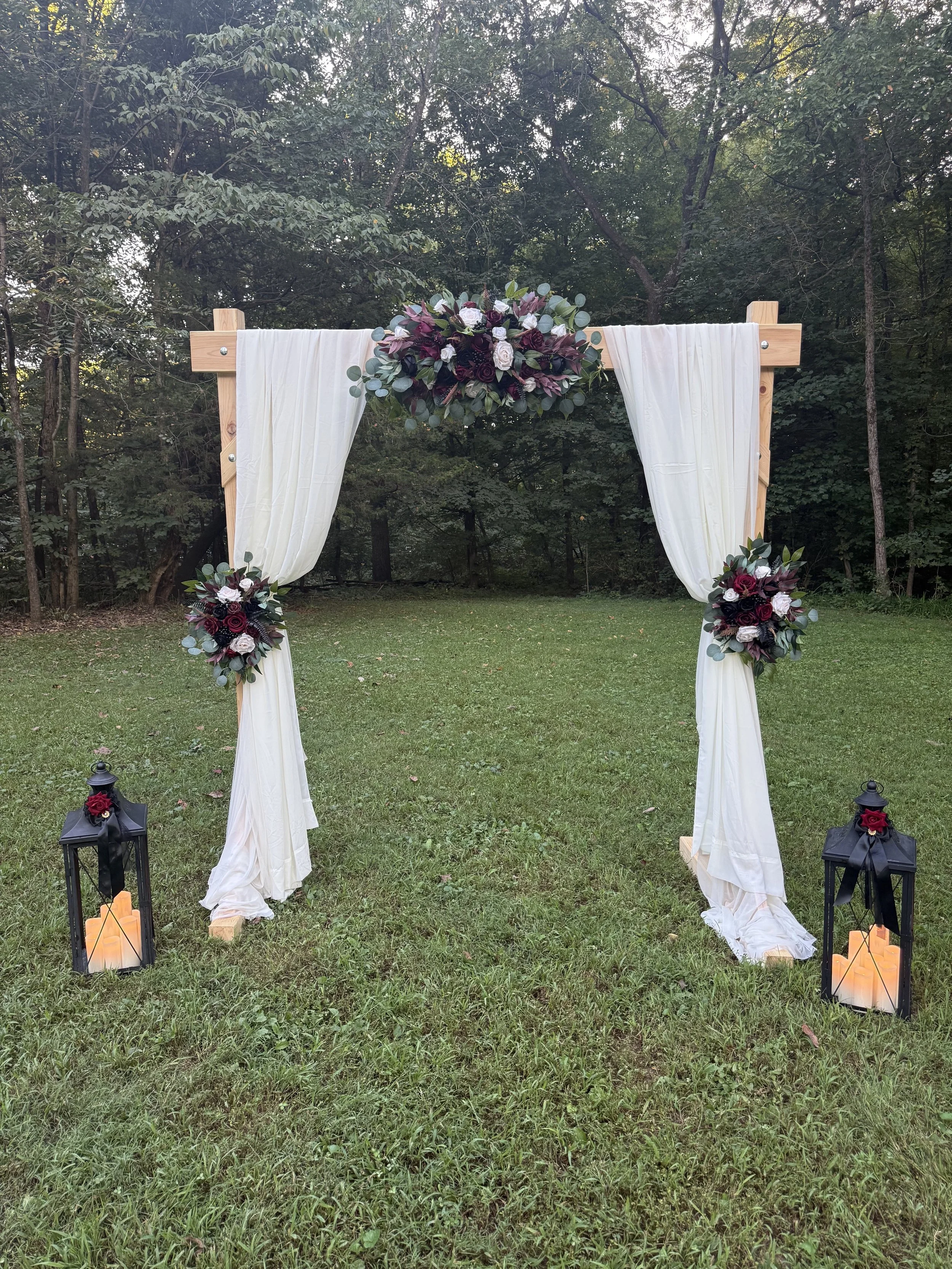 A wedding arch decorated with white curtains, dark purple, white, and green floral arrangements, and positioned on a grassy outdoor area with trees in the background. Lanterns and candles are placed at the base of the arch, creating a romantic settin