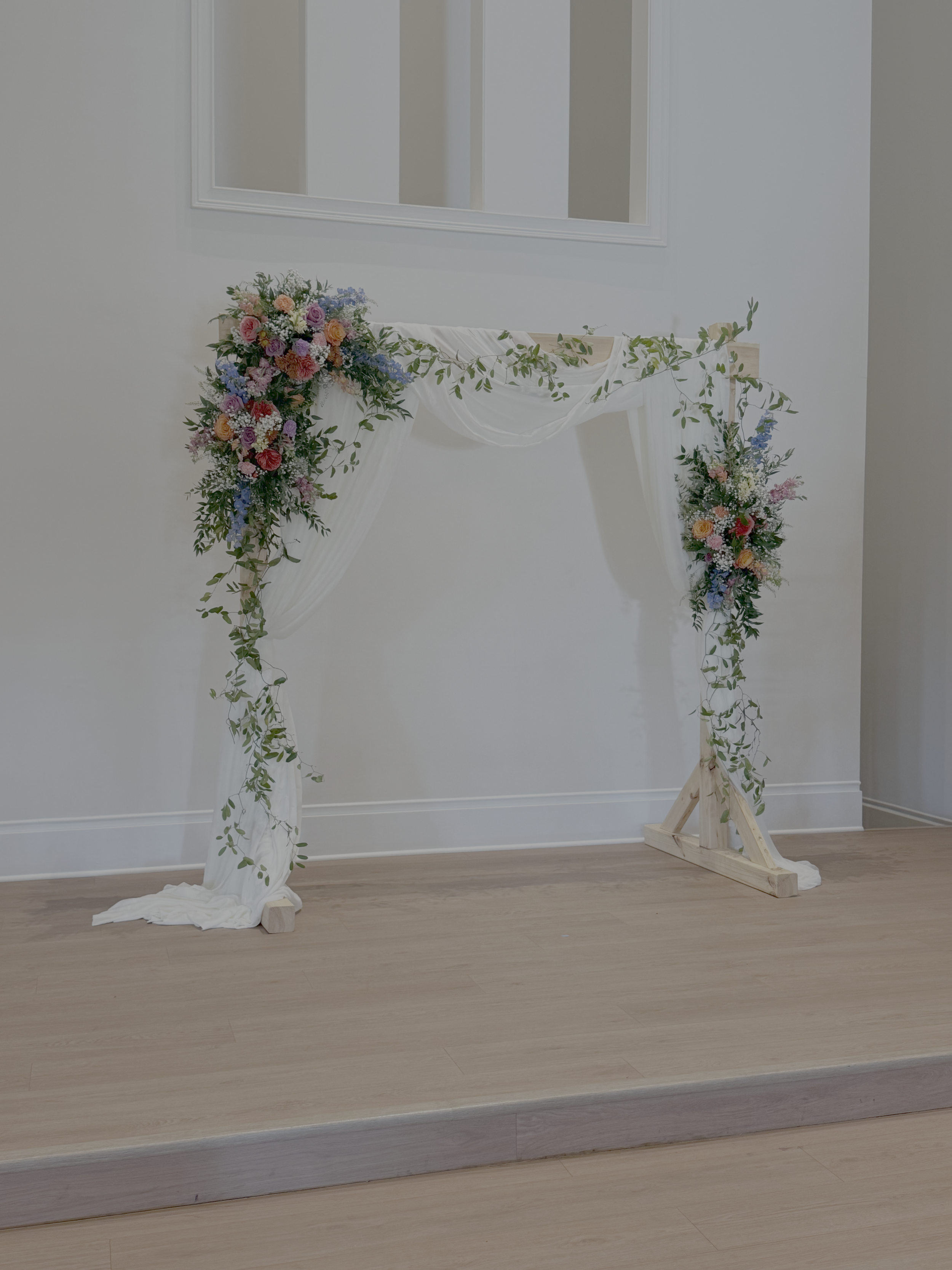 A wedding arch decorated with white fabric and colorful flowers including roses and foliage, set against a plain wall with a large mirror.
