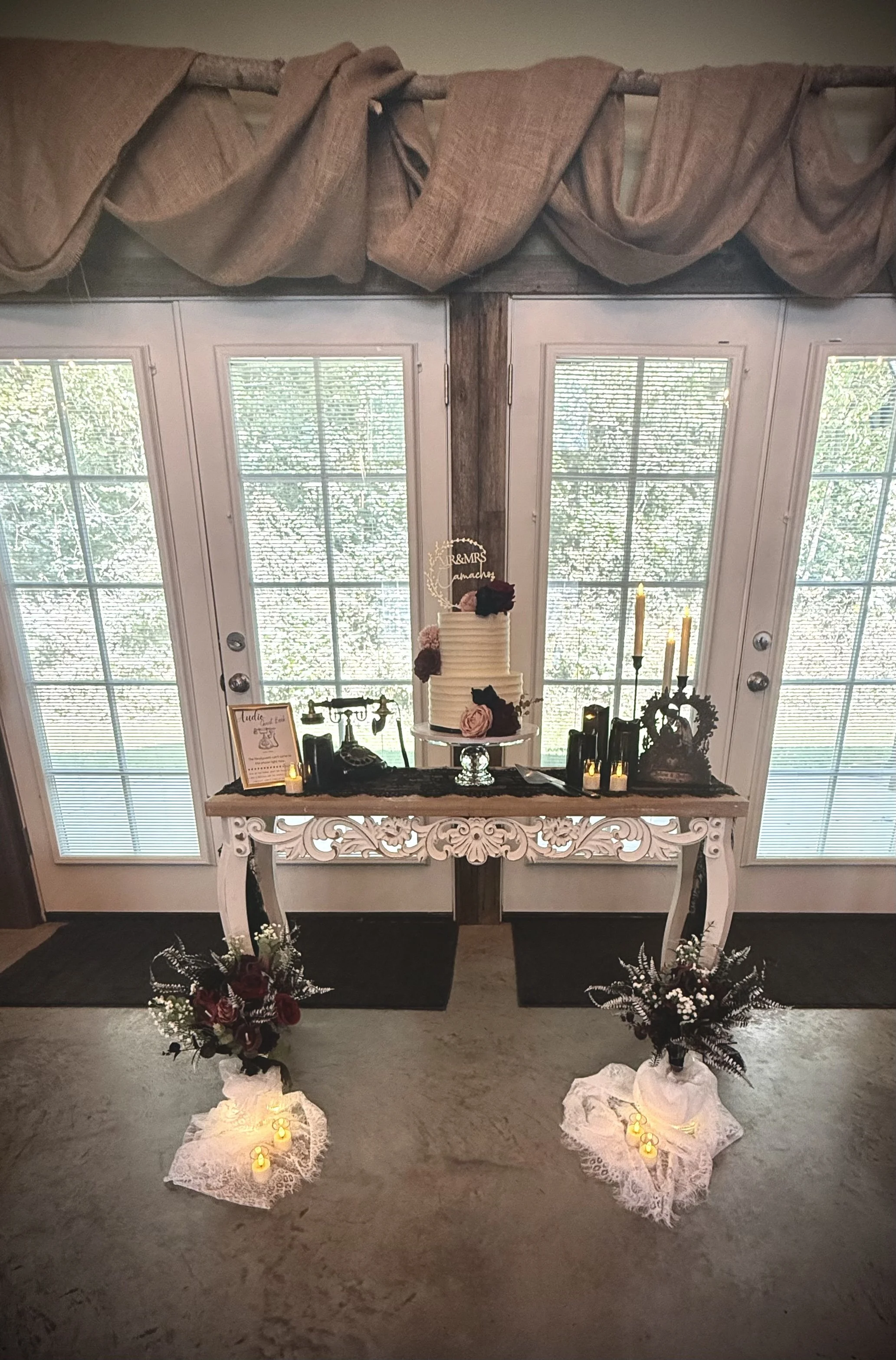 A decorated table setup for a wedding or celebration with a three-tiered cake, floral arrangements, candles, and signs, positioned in front of glass double doors with curtain drapes above.