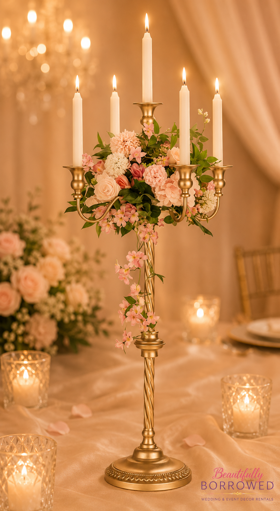 A gold candelabrum with five lit white candles, decorated with pink and white flowers and green foliage, on a table with candlelit glasses and rose petals in the background.
