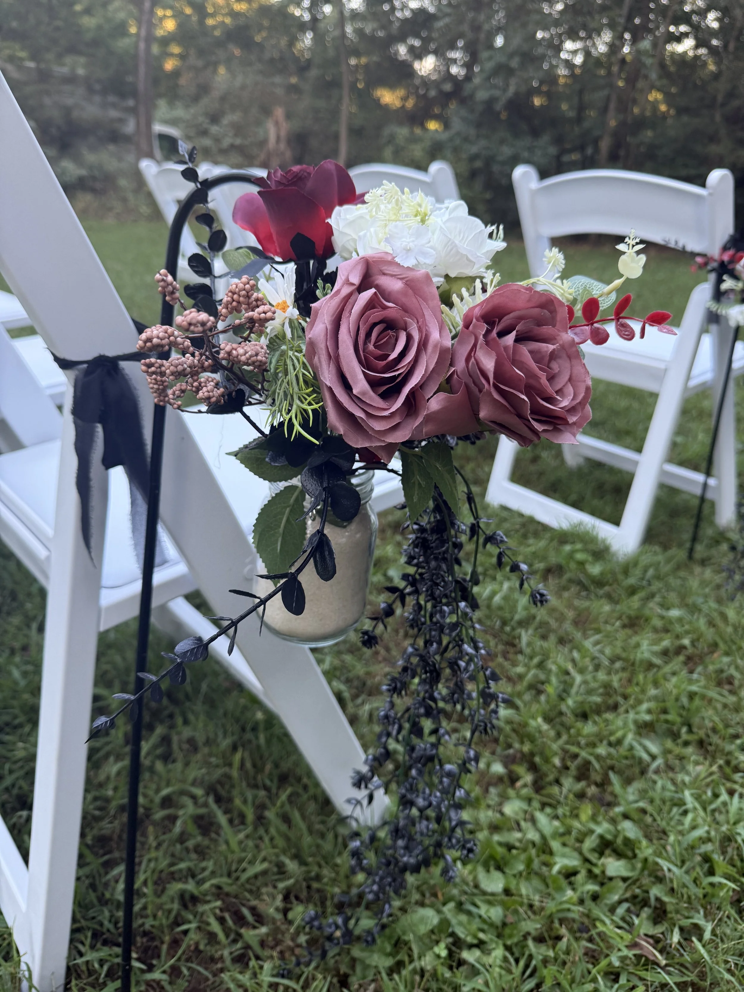 A bouquet of pink, white, and dark red artificial flowers and greenery attached to the side of a white table and chairs outdoors, with a grassy area and trees in the background.
