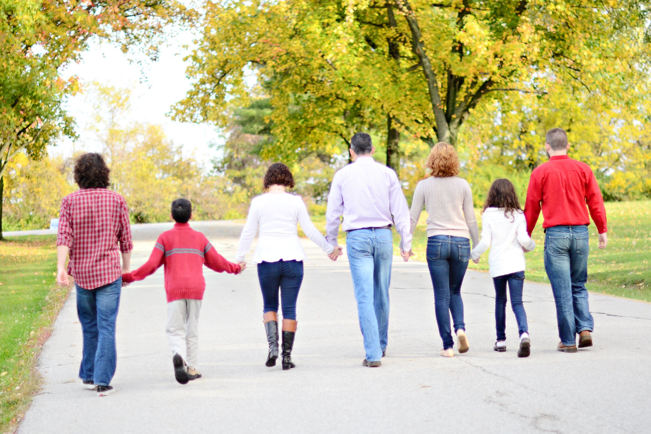 A diverse group of people, including children and adults, walking hand-in-hand on a paved path through a park with trees showing fall foliage.