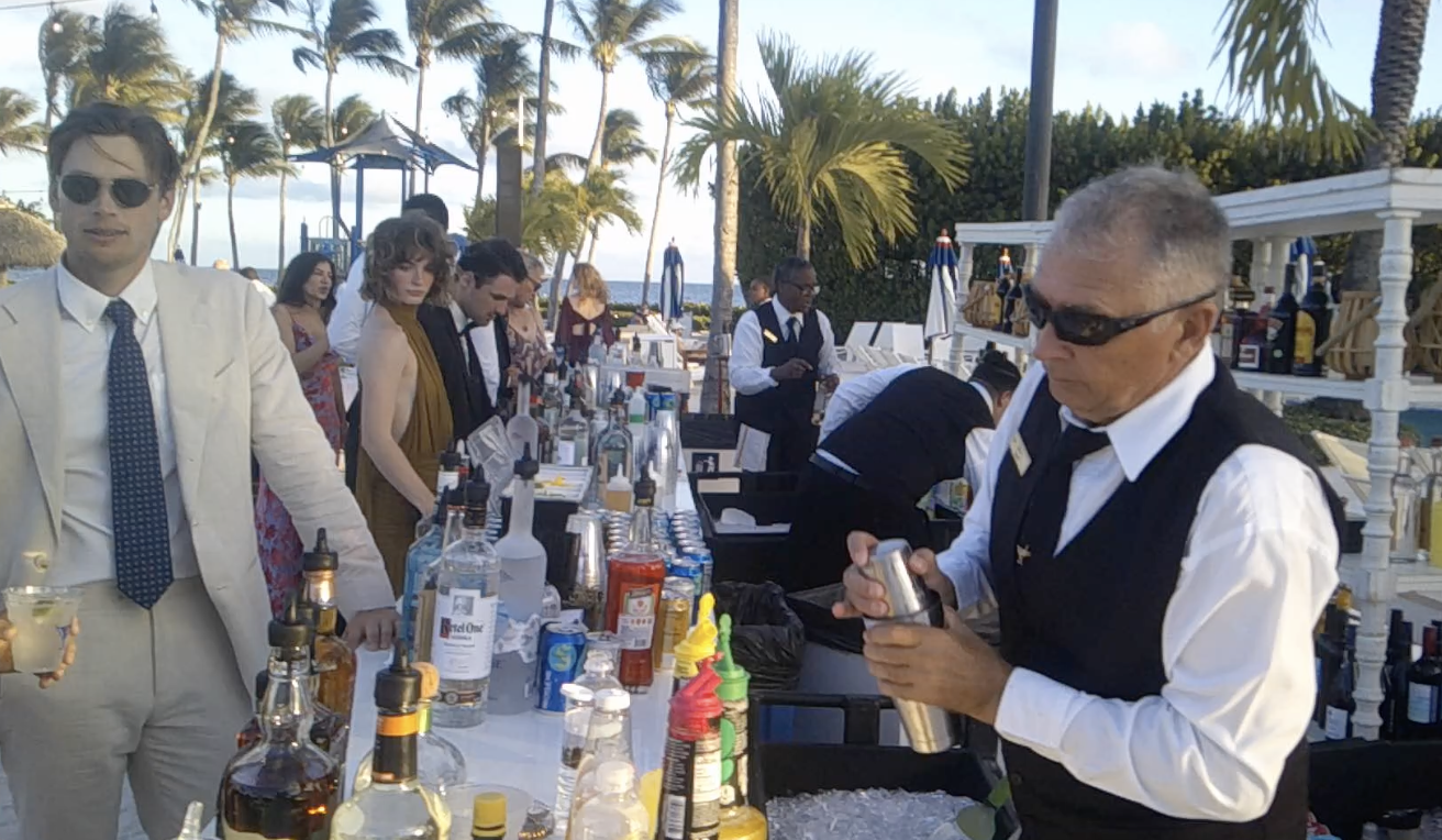People at an outdoor bar or cocktail station on a beachside or tropical resort setting, with bartenders preparing drinks and guests in formal attire enjoying the event.