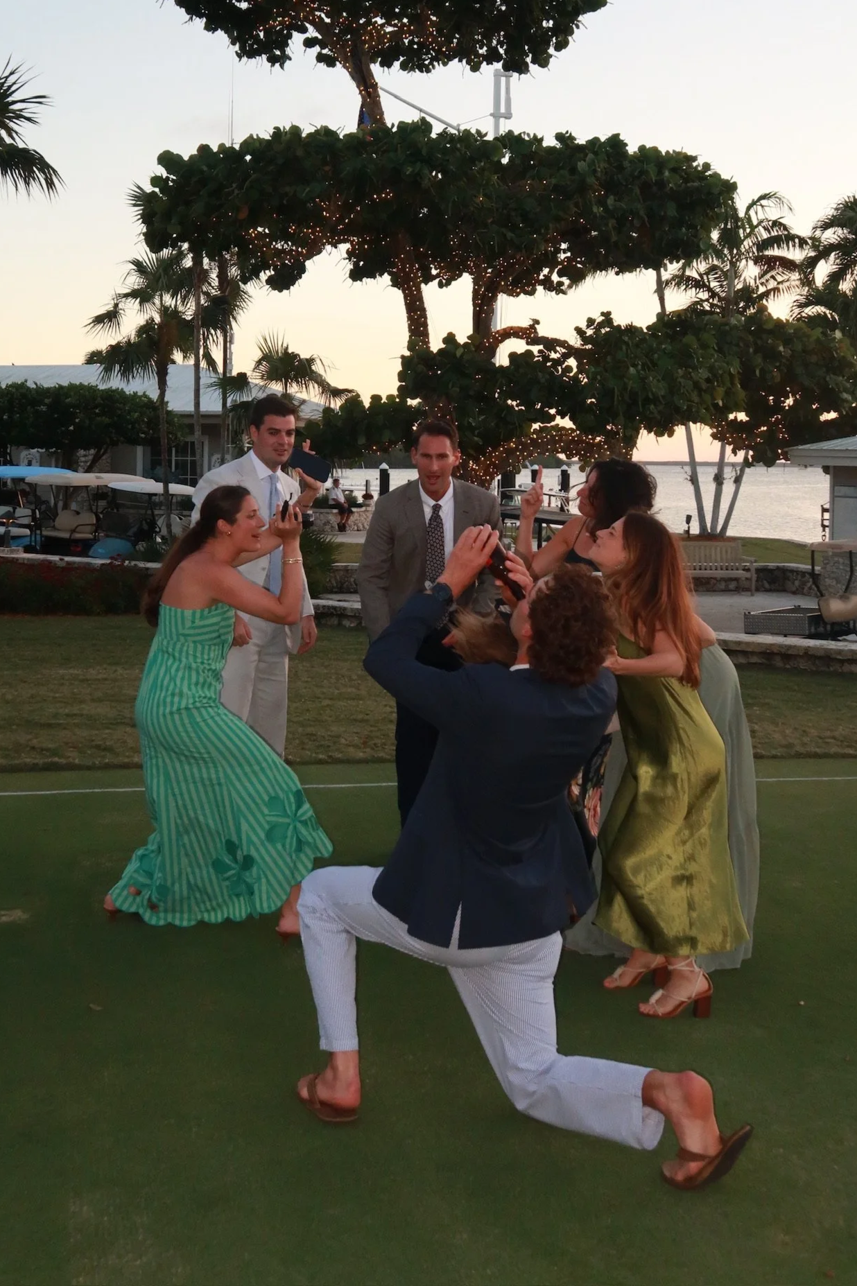 A group of people celebrating outdoors near a lake at sunset, with a large decorated tree in the background. The group includes a woman in a green dress, a man in a gray suit, others in colorful dresses and suits, with some holding drinks and taking pictures.
