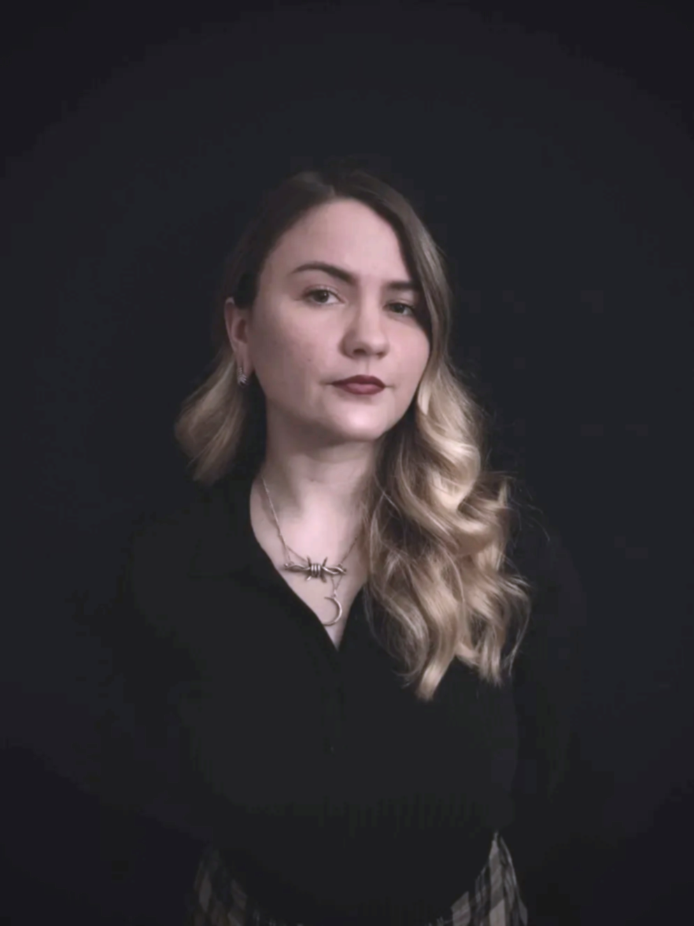 A woman with blond wavy hair, wearing a black top with a necklace, standing against a black background.