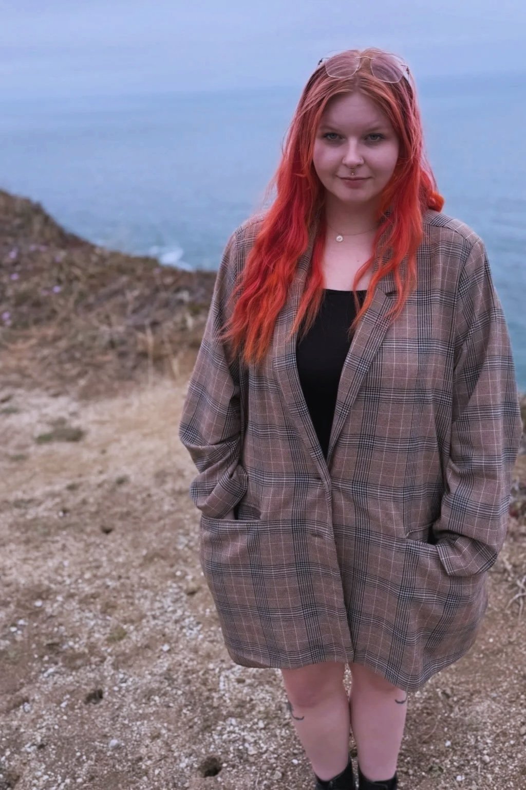 A young woman with red hair, wearing a plaid blazer over a black top, stands on a dirt path near the coast with the ocean in the background.
