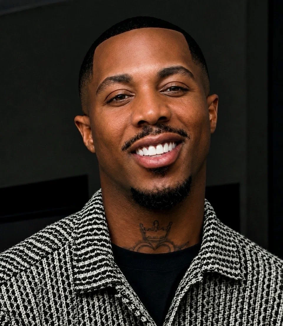 Close-up of a smiling Black man with a goatee and short hair, wearing a patterned jacket, standing against a dark background.