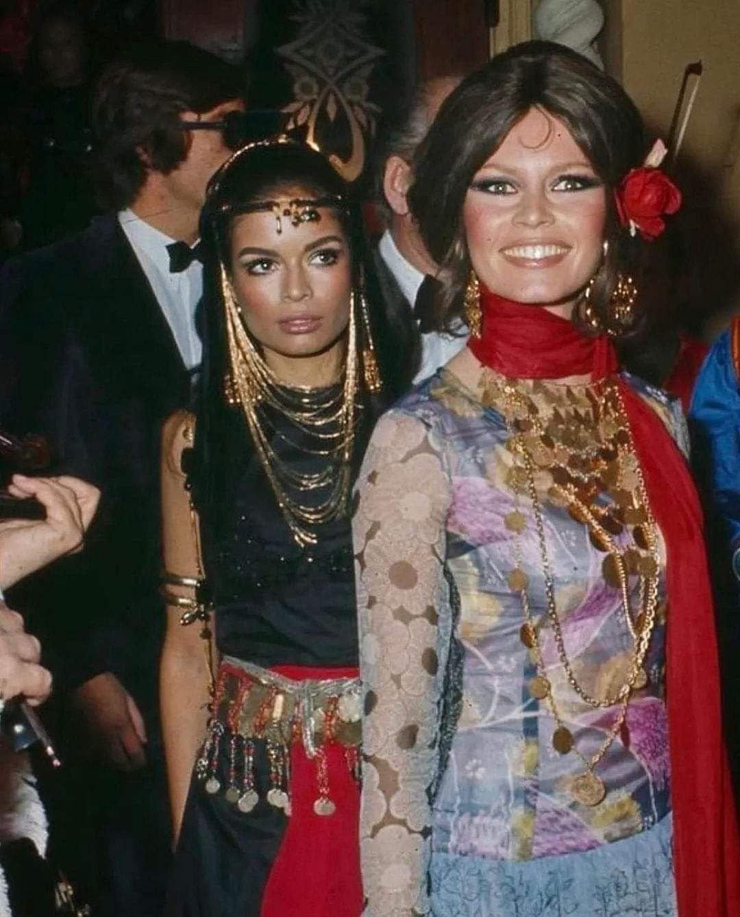 Bianca Jagger and another woman dressed in colorful, ornate costumes with gold jewelry, one smiling and one serious, at a social event.
