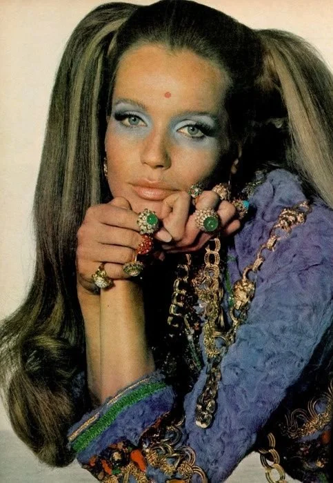 A woman with long styled hair, wearing multiple rings and jewelry, dressed in a colorful, ornate blouse, and has blue eyeshadow with a small red dot on her forehead.
