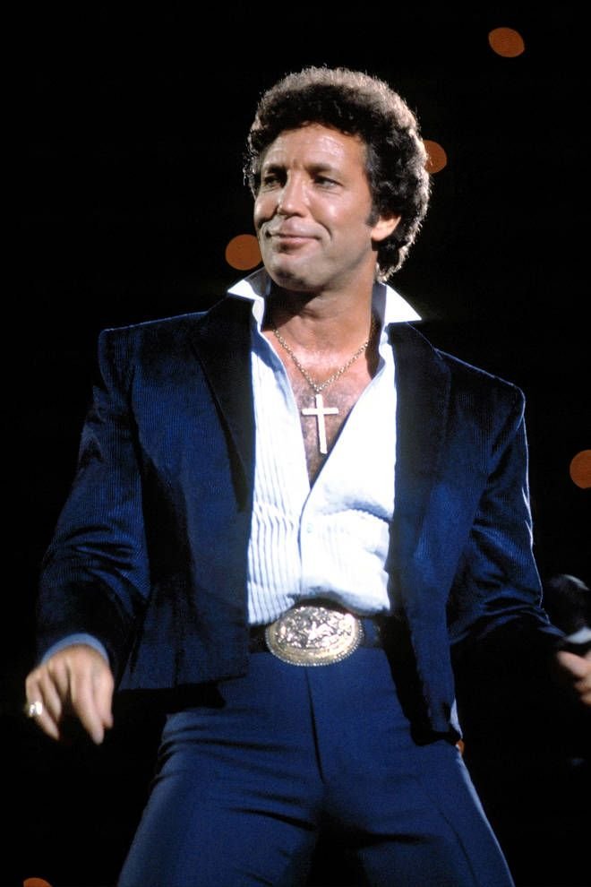 A man in a blue velvet jacket and white shirt, wearing a cross necklace, on stage with blurred lights in the background.