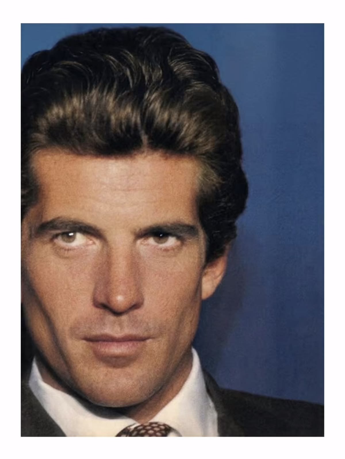 Close-up of JFK Jr. with dark, styled hair, dressed in a suit, against a dark background.