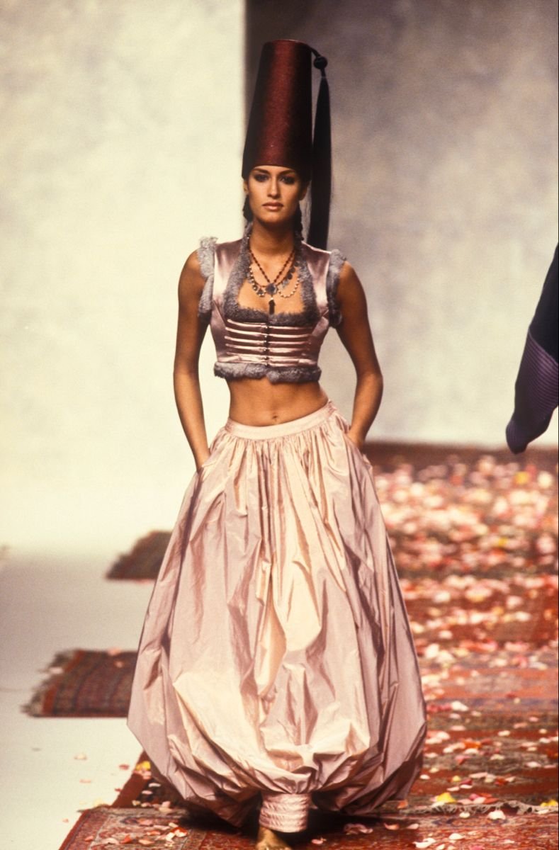 Fashion model walking down runway in an eccentric outfit with a tall headpiece, cropped top, and voluminous skirt.