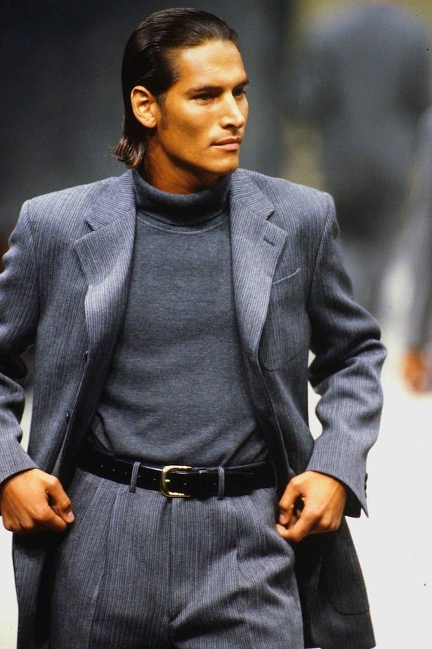 A man with long dark hair dressed in a gray pinstripe suit and turtleneck, standing confidently with hands on hips.