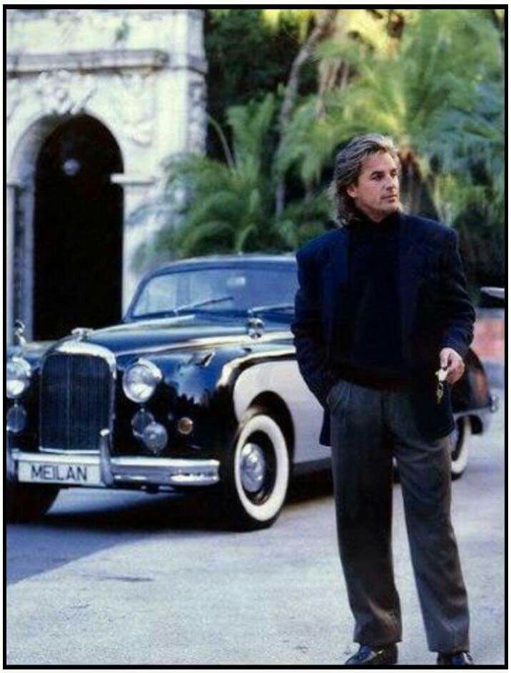 Don Johnson dressed in a black turtleneck, blazer, and gray pants holds keys and stands on a street next to a vintage black car with whitewall tires and a European-style license plate that reads 'MELAN'. 
