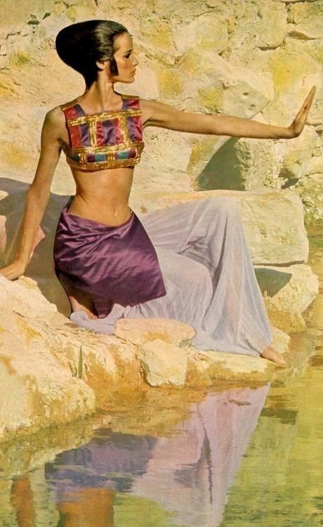 A woman with an elaborate hairstyle sitting on rocks by water, wearing a colorful top and flowing skirt, reaching out her arm toward the water.