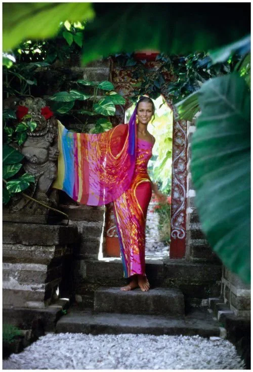 Lauren Hutton in a colorful, patterned dress standing in a lush, tropical garden doorway surrounded by large green leaves and decorative plants.