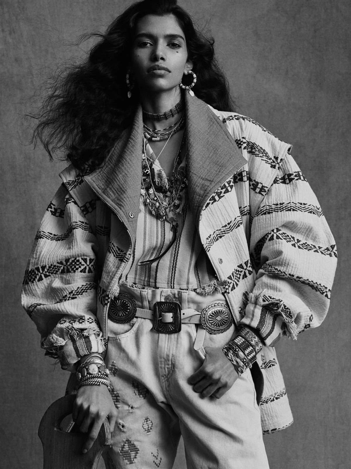 A woman wearing a patterned jacket, layered jewelry, and high-waisted pants poses against a plain background, in black and white.