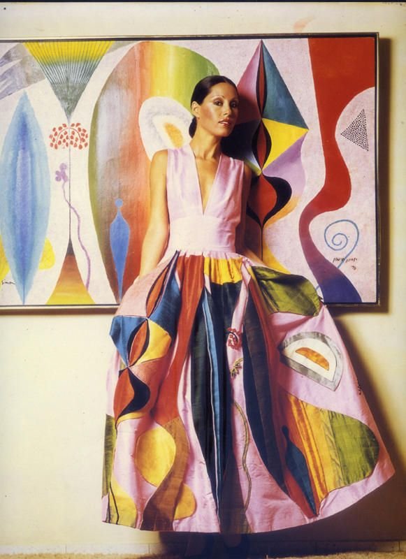 Woman standing in front of a colorful abstract painting, wearing a pink sleeveless top and a voluminous skirt with bold, abstract patterns.