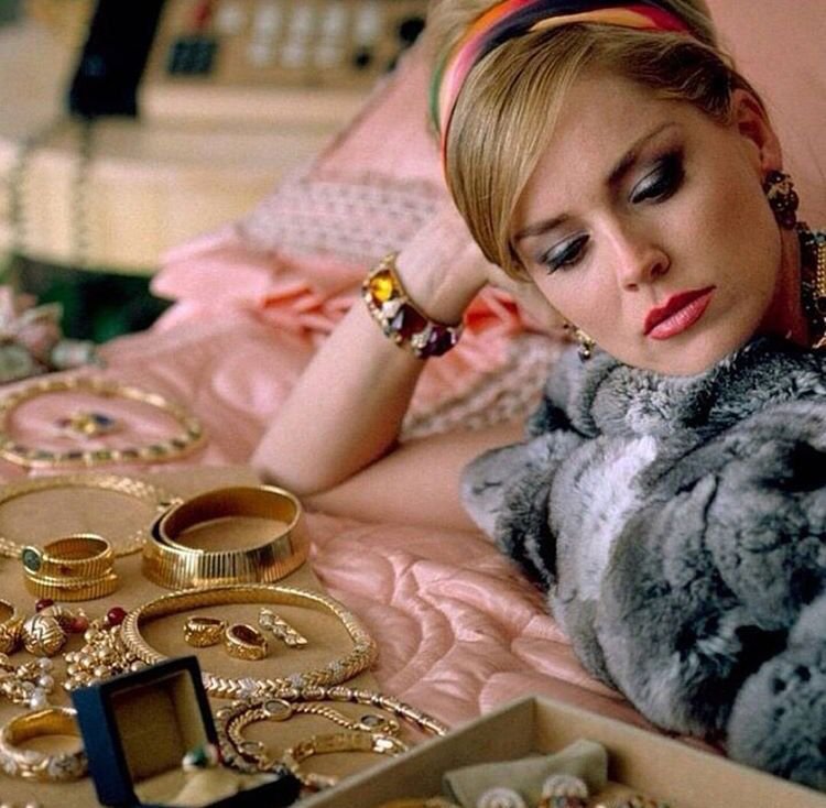 Sharon Stone as Ginger in Casino lying on a pink satin bed surrounded by gold jewelry, including bracelets, rings, earrings, and necklaces, with makeup and a colorful headband.