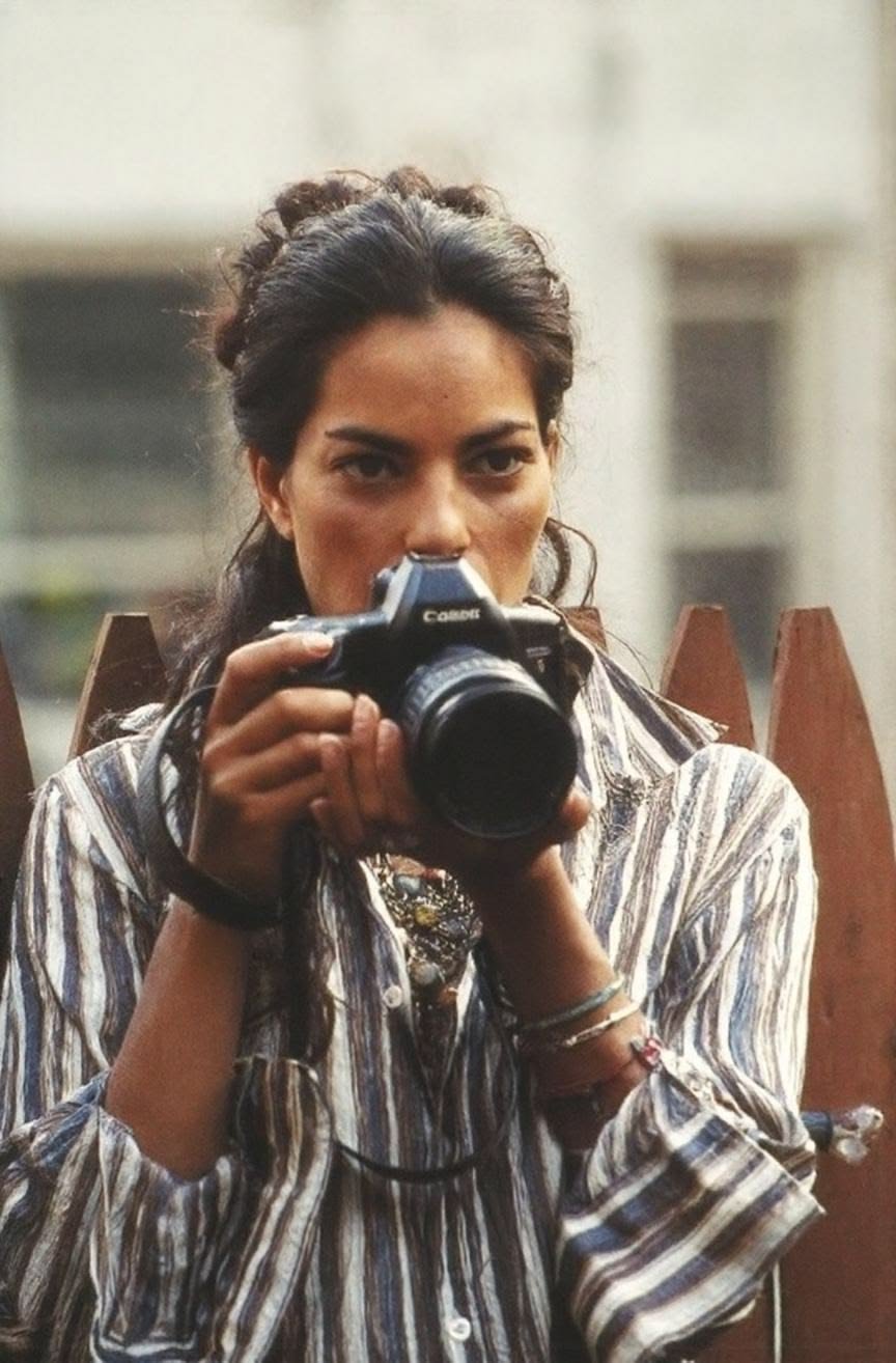 Sarita Choudhury taking a photo with a Canon camera, wearing a striped shirt, with a serious expression, in an outdoor urban setting.