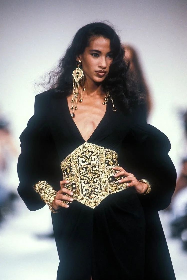 A woman with dark, curly hair modeling a black dress with gold embroidery and jewelry at a fashion show.