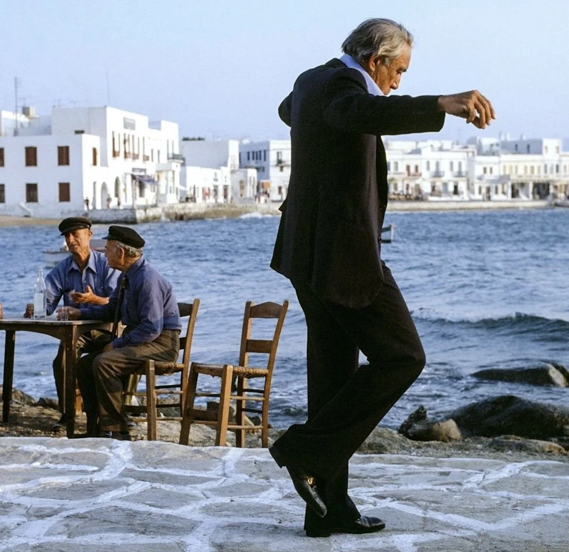 An older man in a black suit dancing on a stone pavement near the water, with two men sitting at a table in the background and white buildings along the shoreline.