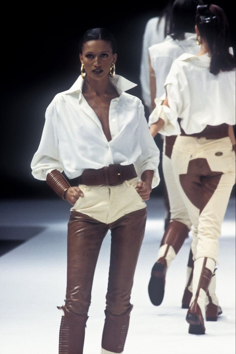 Model walking on runway wearing white blouse and two-tone brown and beige pants with large hoop earrings and cuff bracelet.