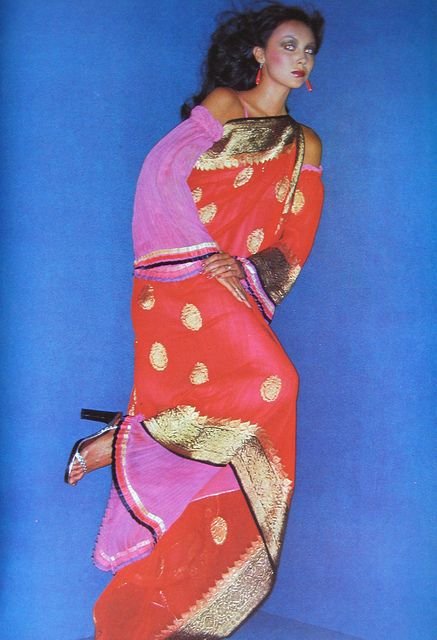 A woman wearing a colorful, traditional-style sari with gold embroidery and a pink shawl posing against a blue background.