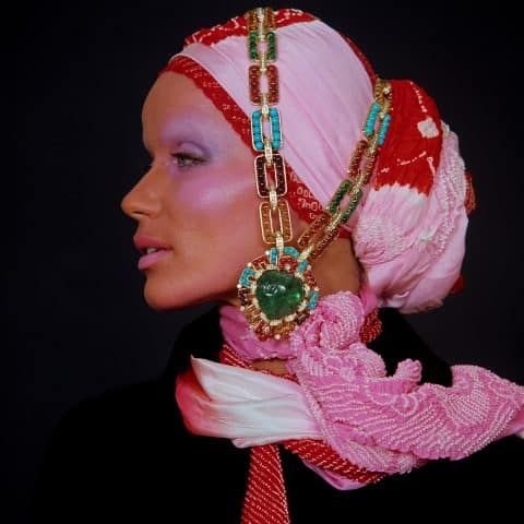 Profile of a woman wearing a pink and red patterned headscarf adorned with large, colorful jewelry and a black outfit with a red and white scarf against a dark background.