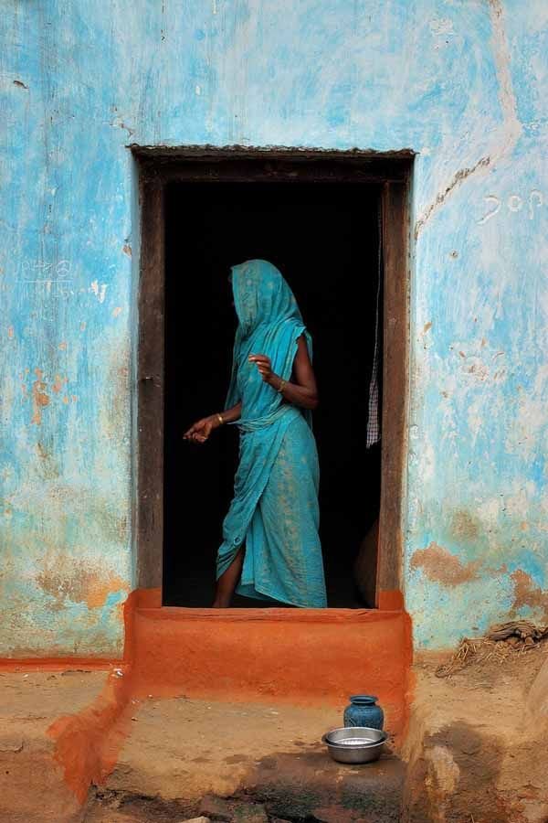 A woman in a blue sari standing in the doorway of a weathered blue building with a dark interior.