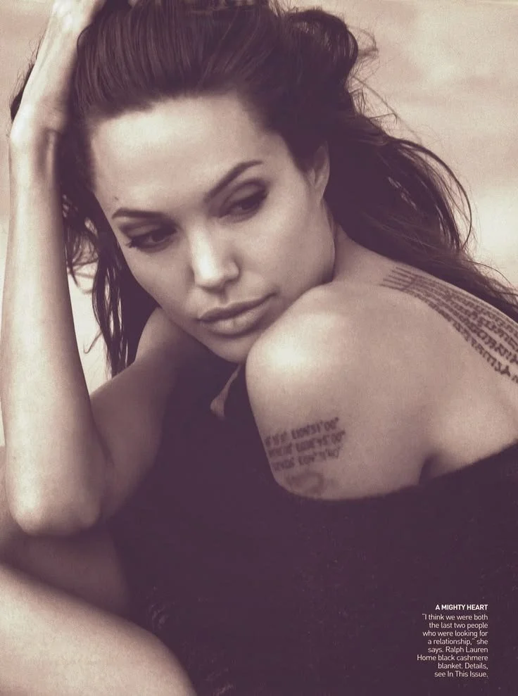 Angelina Jolie lying down, resting her head on her hand, looking thoughtfully at the camera. She has tattoos on her shoulder with text.