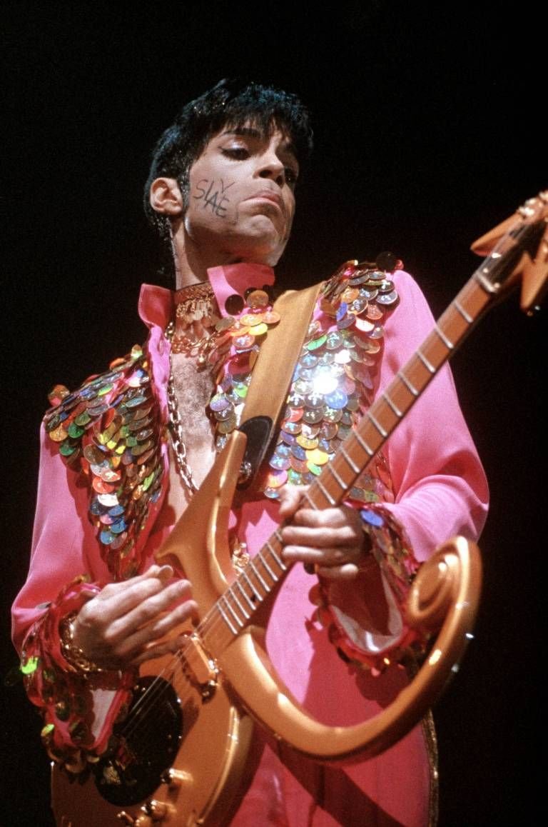 Prince dressed in a vibrant pink outfit with sequin embellishments, playing an electric guitar on stage.