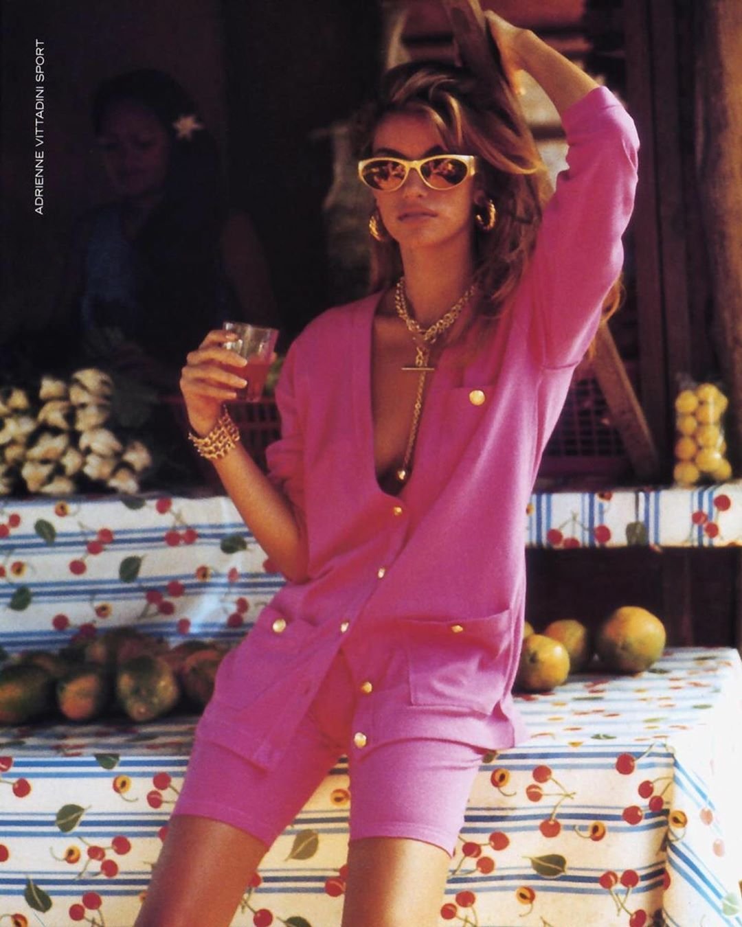A woman with wavy blonde hair, wearing pink sunglasses, a pink blazer, and matching shorts, holds a drink in her left hand, standing in front of a fruit stand with apples and lemons, in a colorful setting.
