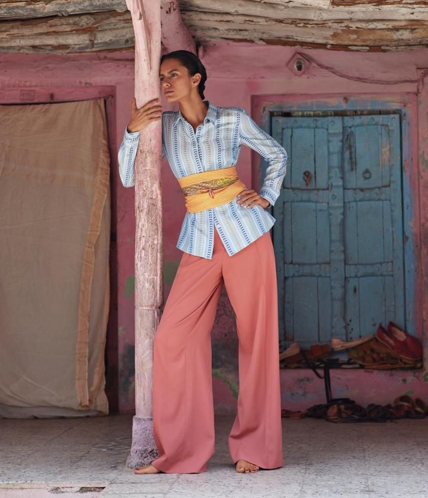 Woman standing outdoors leaning against a pink-painted wooden post, wearing a striped blue and white shirt, wide-legged pink pants, and a yellow sash, with a pink textured wall and blue shuttered window in the background.