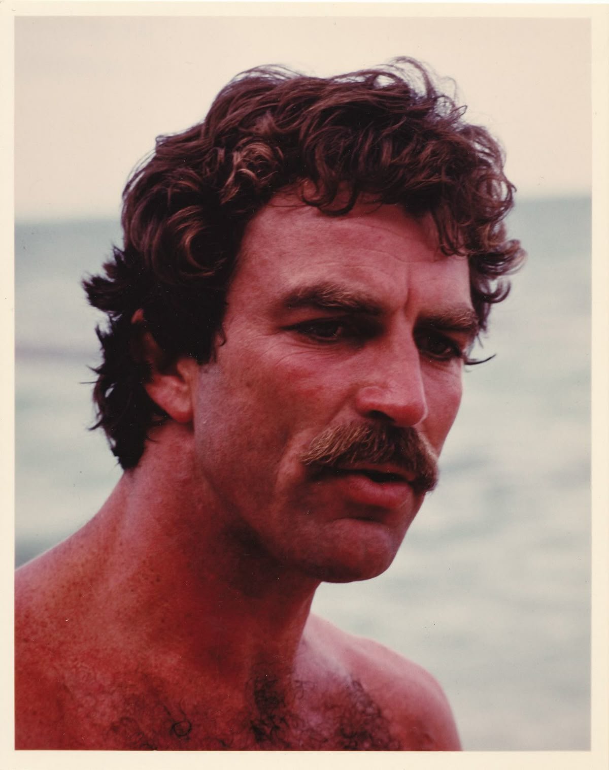 A shirtless man with curly brown hair and a mustache at the beach.