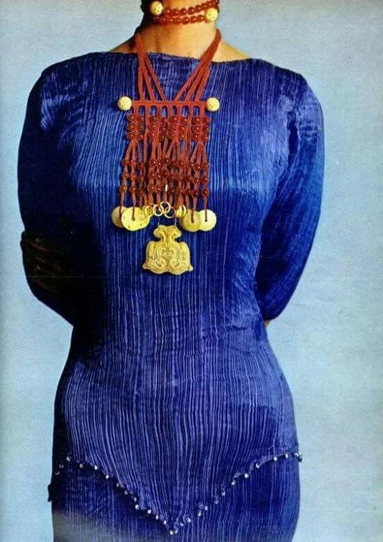 A person wearing a blue ribbed dress with long sleeves, adorned with a red beaded necklace featuring a large golden jewelry piece hanging from it.
