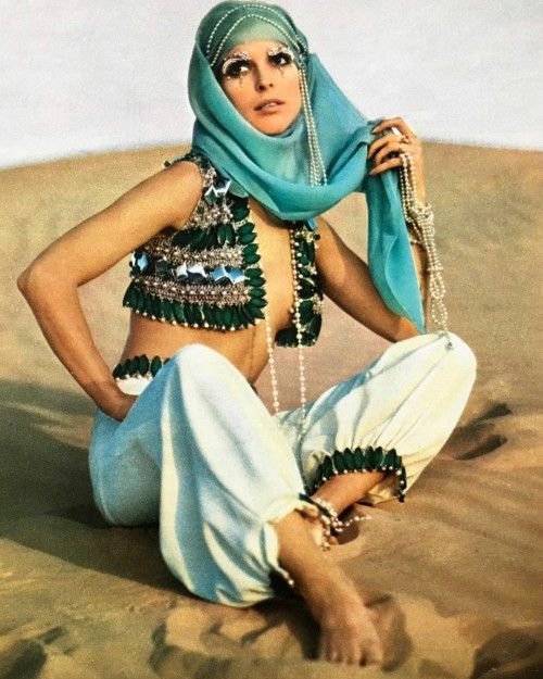 A woman sitting on desert sand, wearing a headscarf, beaded jewelry, and a decorative outfit with traditional elements.